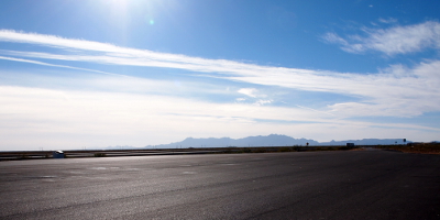 [A long expanse of pavement meets the horizon where a blue haze covers distant mountains. White streaks of clouds stream across the sky while the sun flashes light onto the scene.]